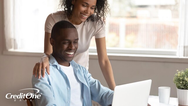 A happy couple smiles as the wife watches her husband working on his laptop. This is how you may react when you redeem rewards from your credit card.