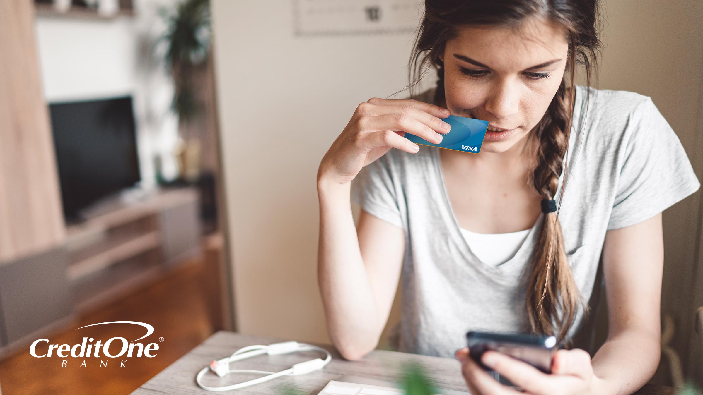 A young woman holds a credit card while looking at her smartphone, appearing thoughtful and possibly wondering if she can reopen a closed credit card.