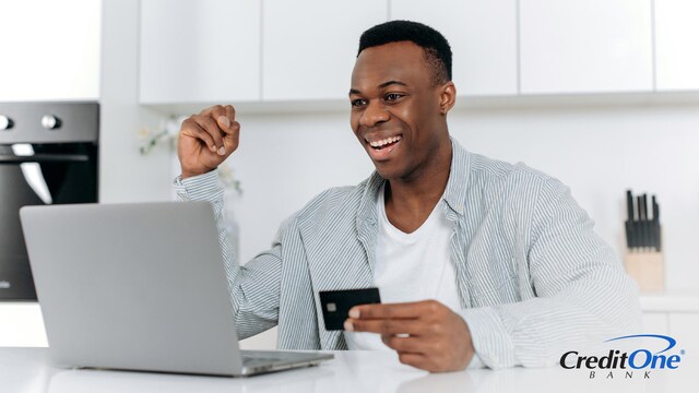 A young man smiles and pumps his fist in excitement as he holds a new credit card while using his laptop.
