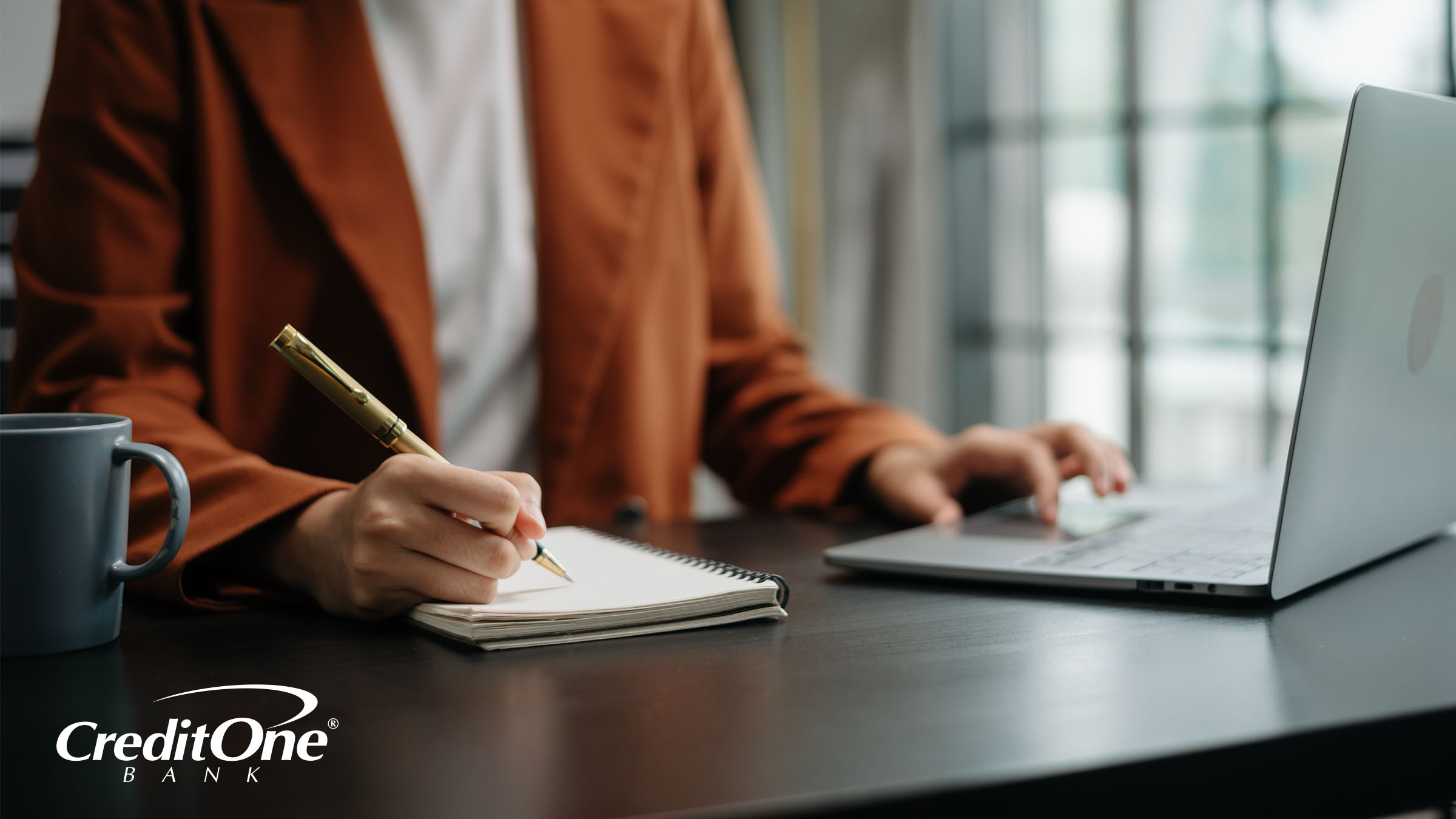 A woman, framed from the neck down, sits at a desk with coffee and a laptop while writing in a spiral notebook. She looks as if she may be checking items off a list, a useful tool for staying on top of credit card payments.