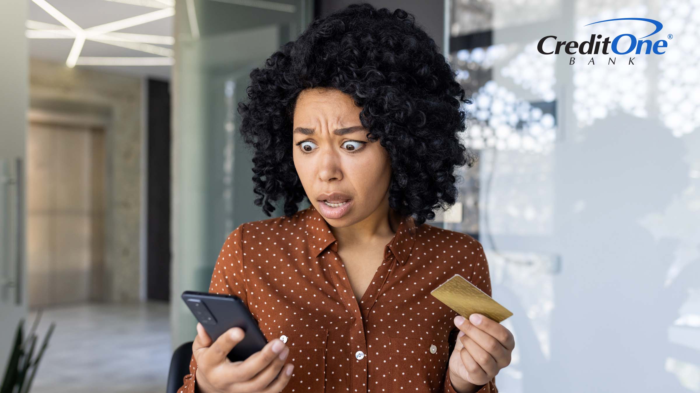 A woman has a shocked expression as she looks at her phone while holding her credit card. She may have fallen prey to one of the latest credit card scams.